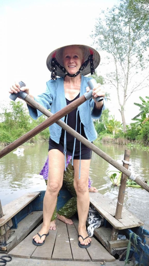 Rowing a wooden boat standing up, in a conical hat.