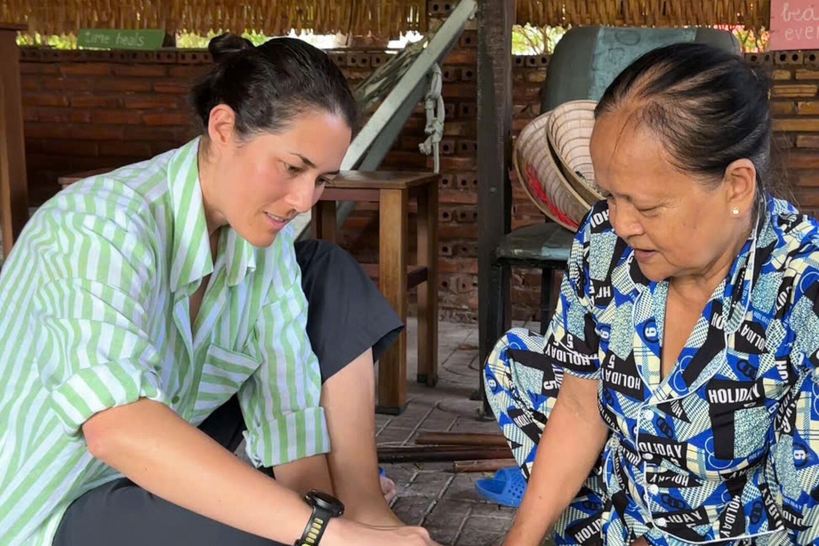 Two women sitting on a floor under a thatched shelter, both looking down at their hands.