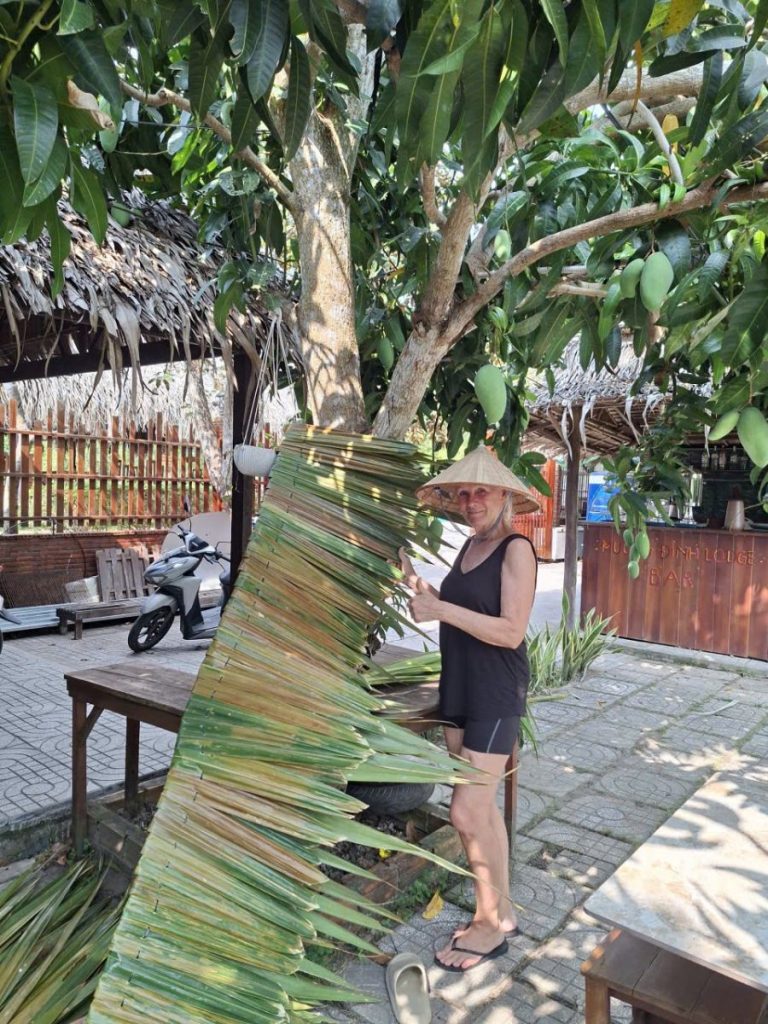 A woman in a conical hat beside long thatch panels propped against a tree.