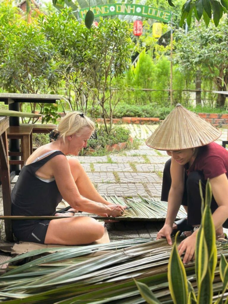 A guest and a woman in a conical hat weaving thatch panels on the ground.