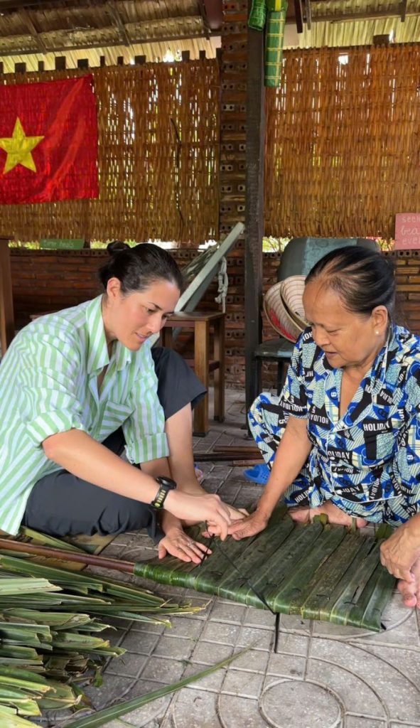 A guest learning to weave thatch from a local woman, a Vietnamese flag on the wall behind.
