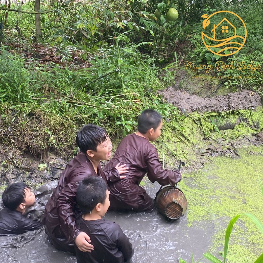 Four boys in brown clothes wading in a muddy ditch, one holding a woven fish basket.