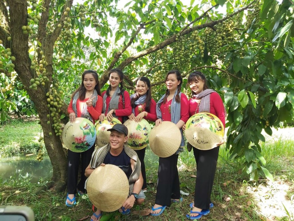 A group in red shirts holding conical hats under a fruit tree in the garden.