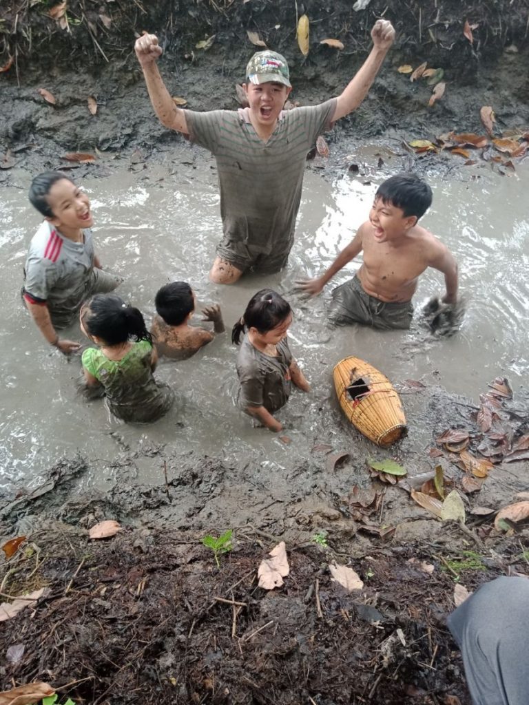 A man cheering with raised arms as children splash in a muddy ditch.