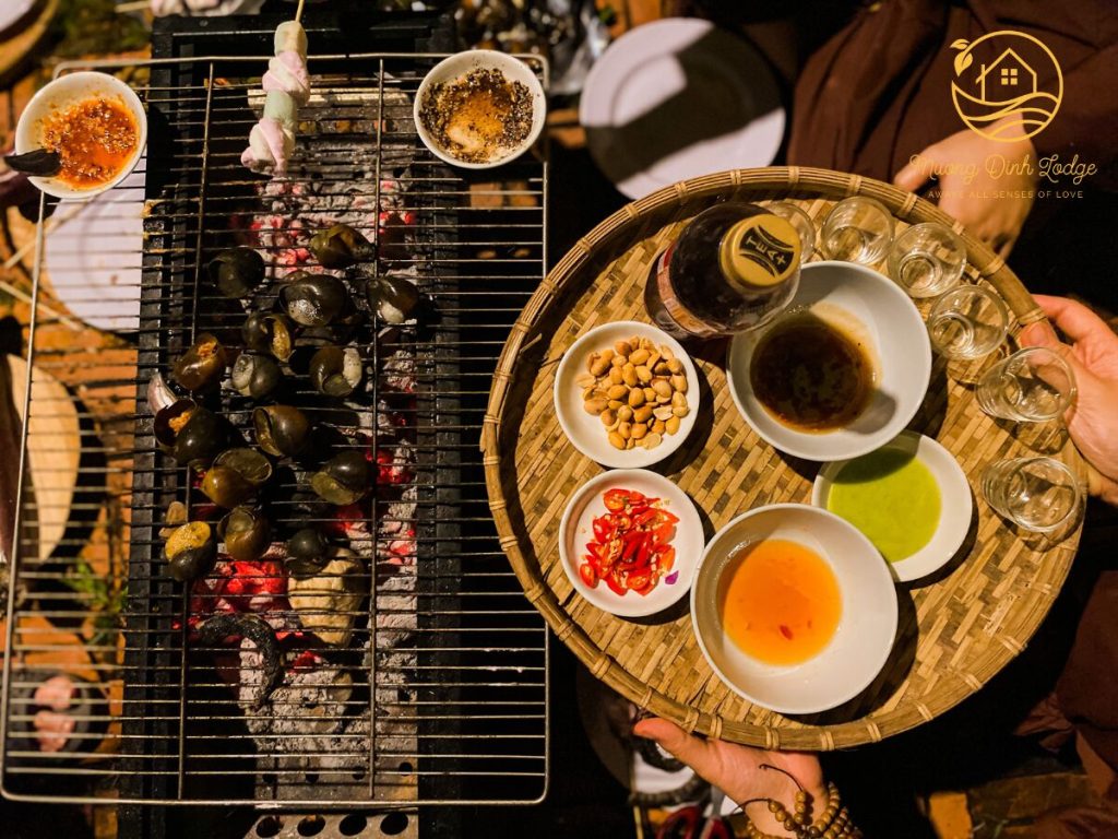 Snails grilling over charcoal beside a tray of dipping sauces and small glasses.