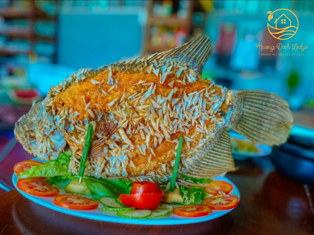 A whole crispy fried fish set upright on a plate with sliced tomato and cucumber.