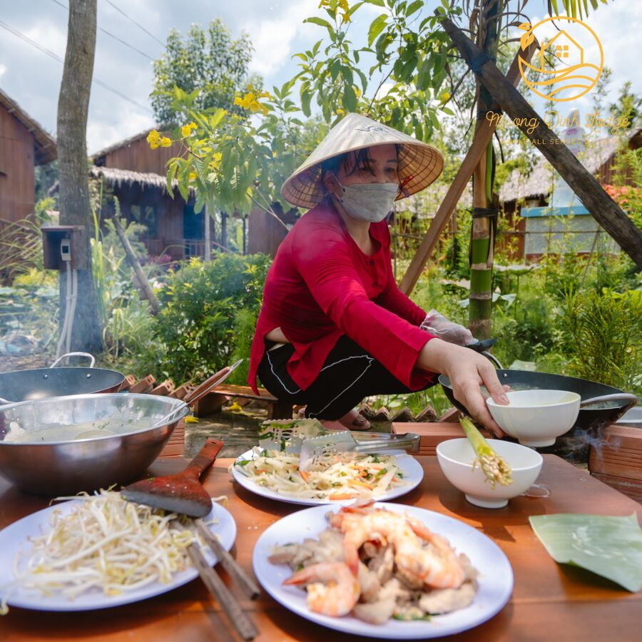 A woman in a conical hat preparing food outdoors beside plates of bean sprouts and prawns.