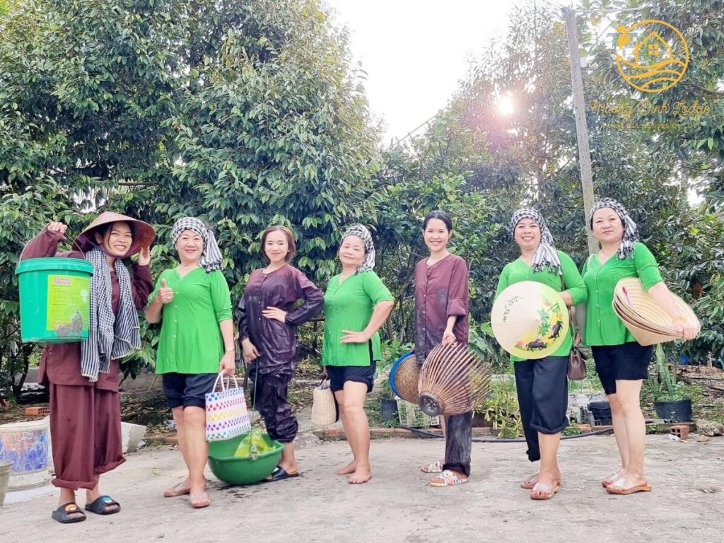 Seven women in green or brown shirts posing in a yard with buckets, bags, fish traps and conical hats.