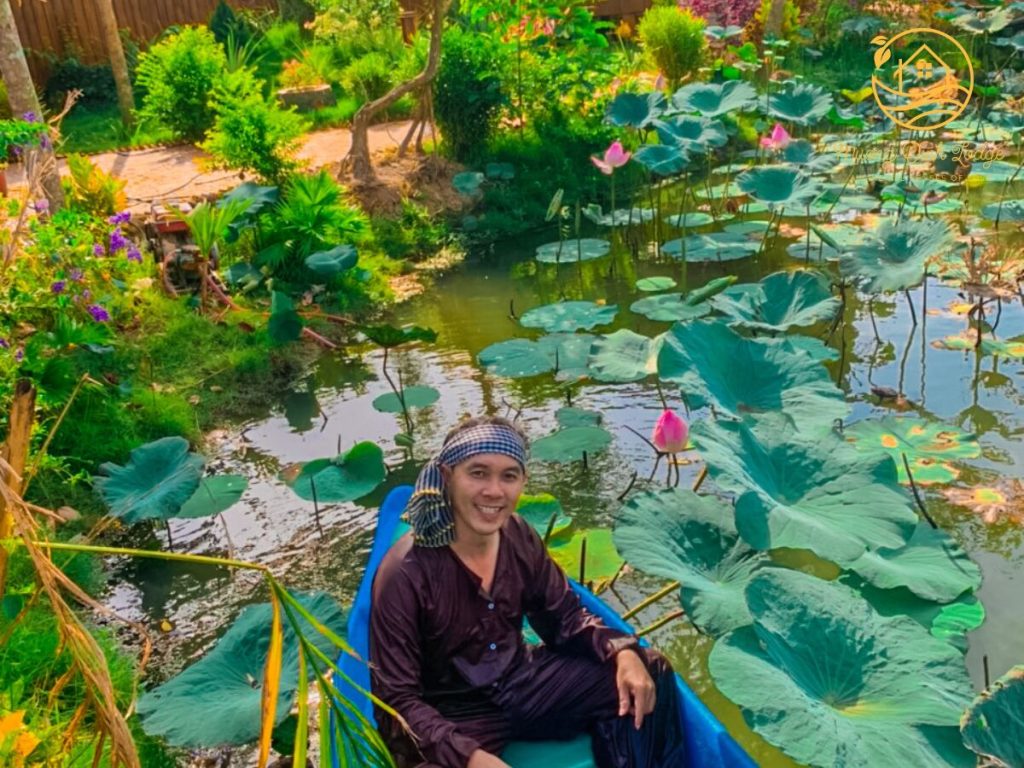 A man in a brown shirt sitting in a small boat among lotus leaves and pink flowers.