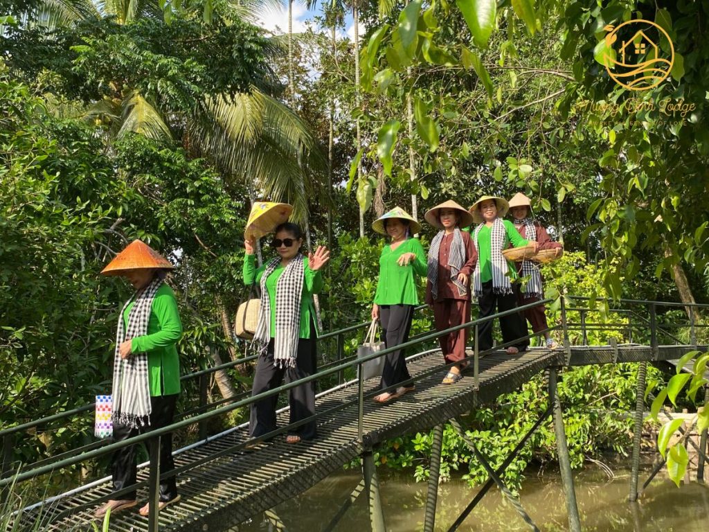 A group in conical hats and checked scarves crossing a narrow footbridge over a canal.