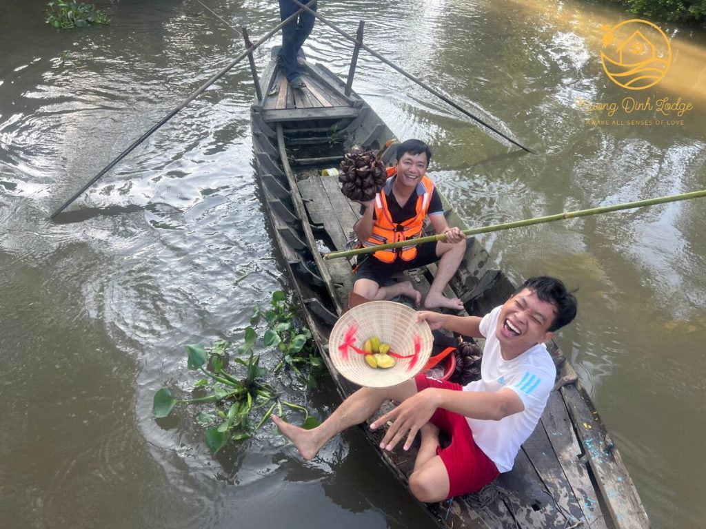 Two laughing men in a wooden boat, one holding fruit in an upturned conical hat, the other a dark fruit cluster.