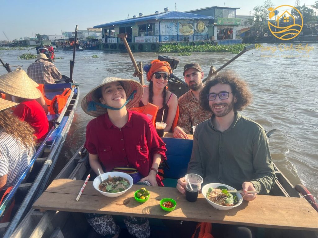 Guests eating bowls of soup at a table set across a boat on a busy river.