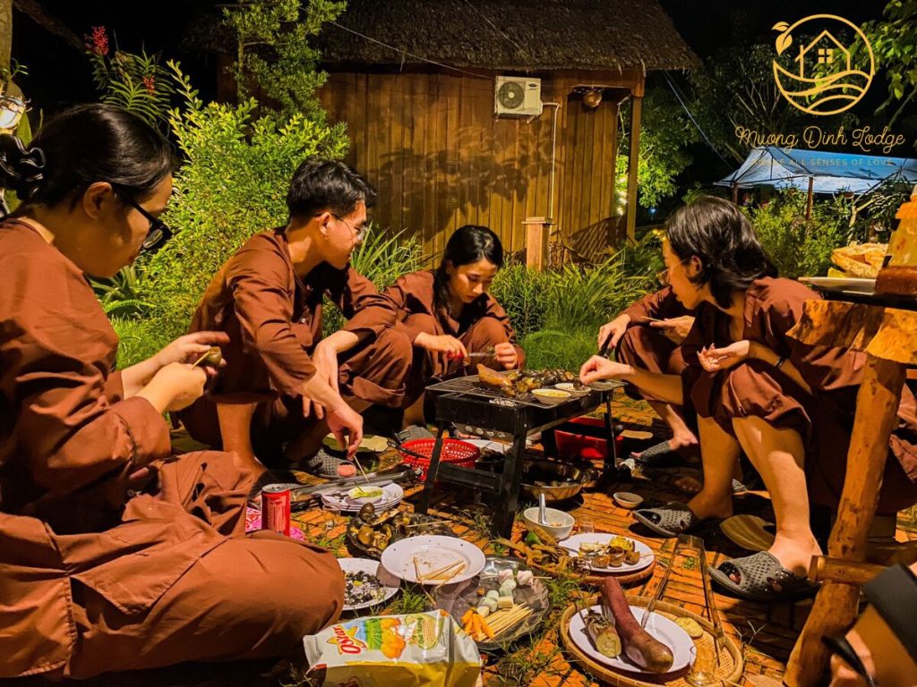 Guests in brown traditional outfits grilling food around a low table at night.