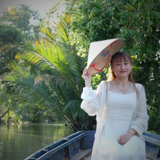 Cô gái mặc váy trắng cầm nón lá ngồi trên xuồng giữa hàng dừa nước.