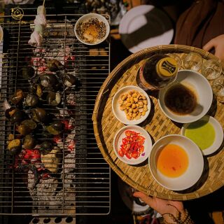 Ốc nướng trên vỉ than cạnh mâm tròn bày các chén nước chấm.