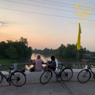 Bicycles parked on a riverside path at sunset while two people sit by the water.