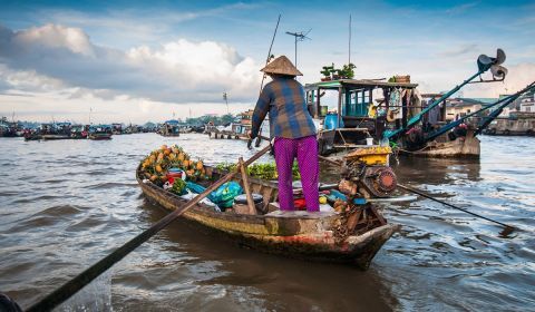A woman in a conical hat rowing a small boat of produce among larger boats on the river.