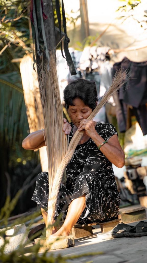 Bà cụ địa phương ngồi bó chổi bằng tay trước hiên nhà.