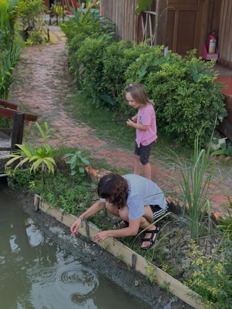 A woman crouching at a canal's edge as a child watches from the garden path.