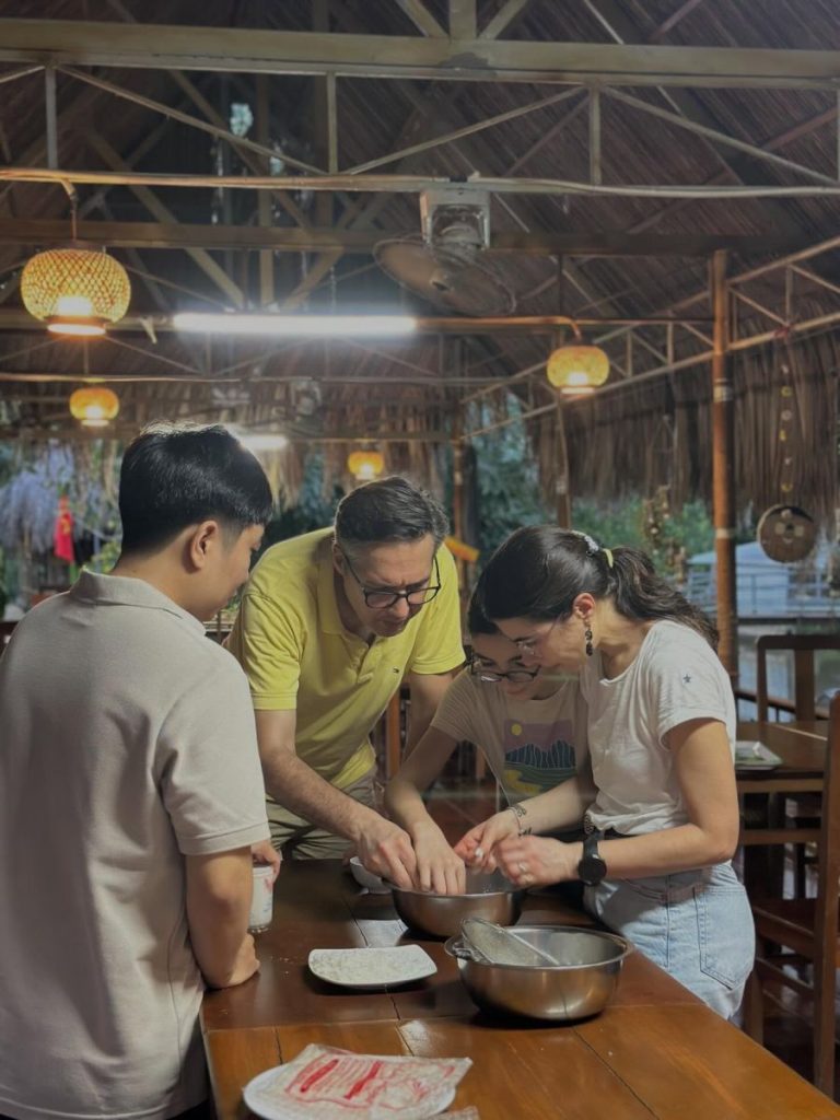 Three people working dough together at a wooden table in an open-air hall.