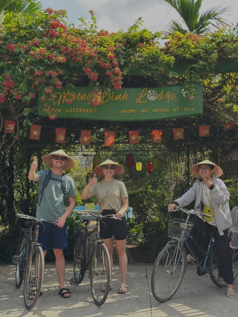 Three cyclists in hats with their bikes at the flower-covered gate of Mương Đình Lodge.