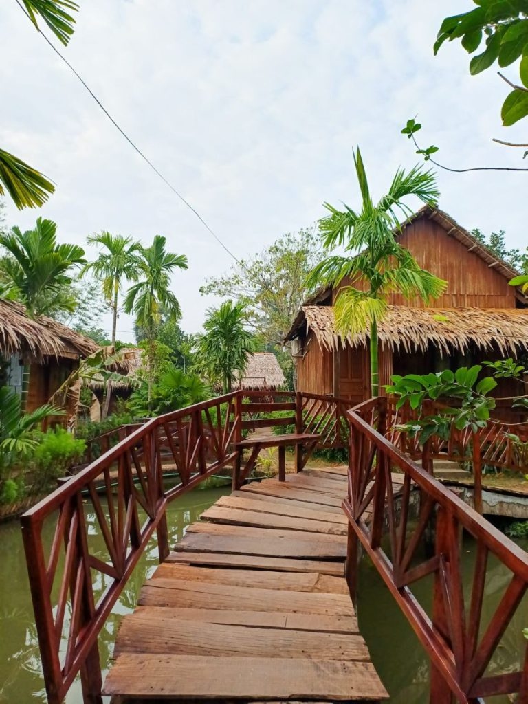 A red-railed wooden footbridge leading toward a thatched cabin among palms.