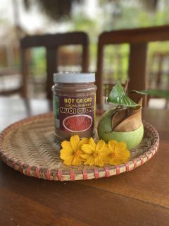 A jar of Mười Cương cocoa powder on a woven tray with green fruit and yellow flowers.