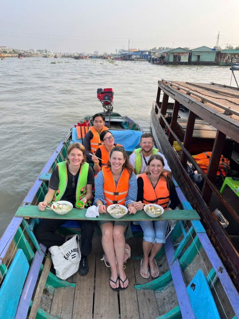 Six smiling people in orange and green life vests at a plank table with bowls of food on a boat.
