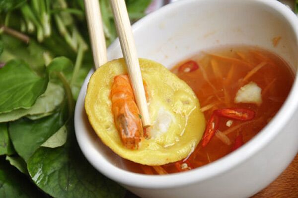Chopsticks dipping a banh khot with prawn into fish sauce, fresh greens beside.