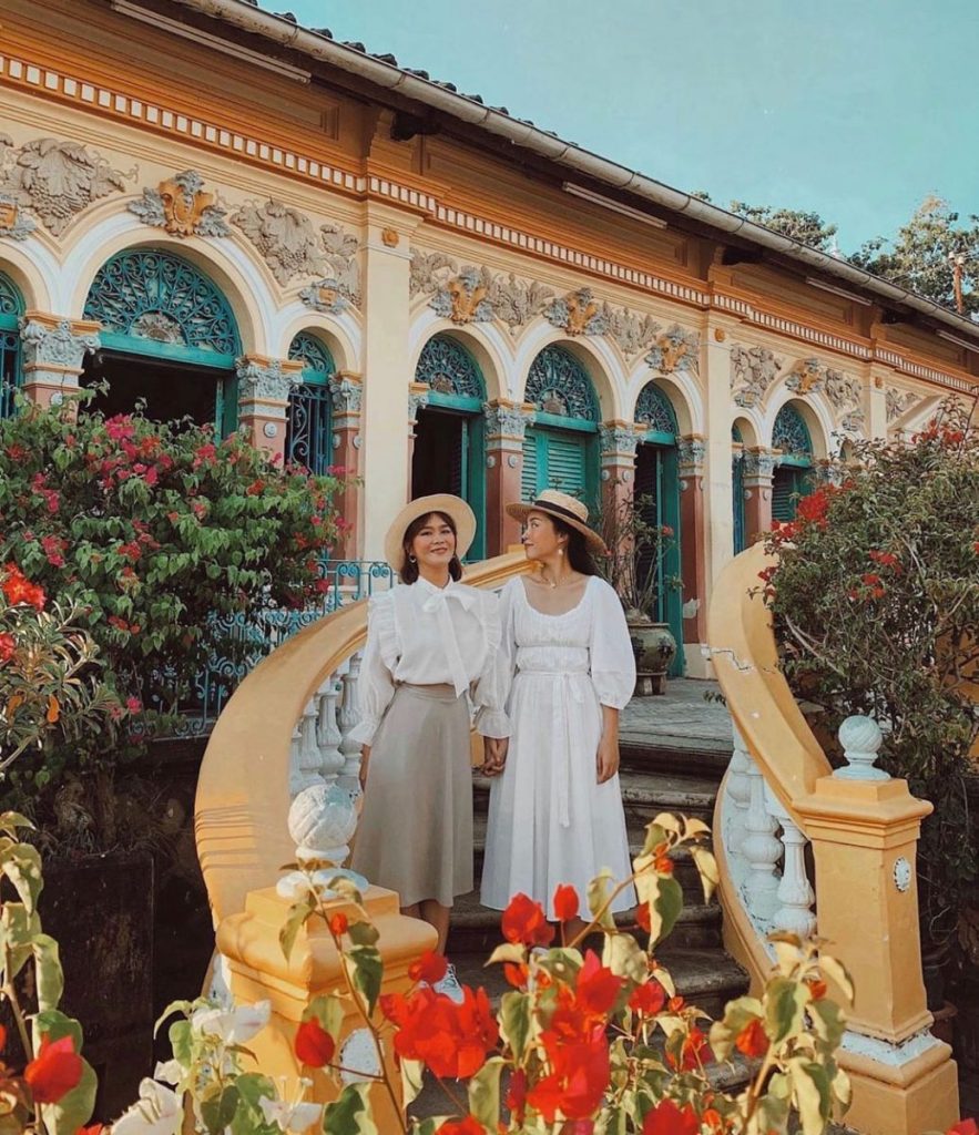 Two women in white on the curved stairs of an ornate old house among red flowers.