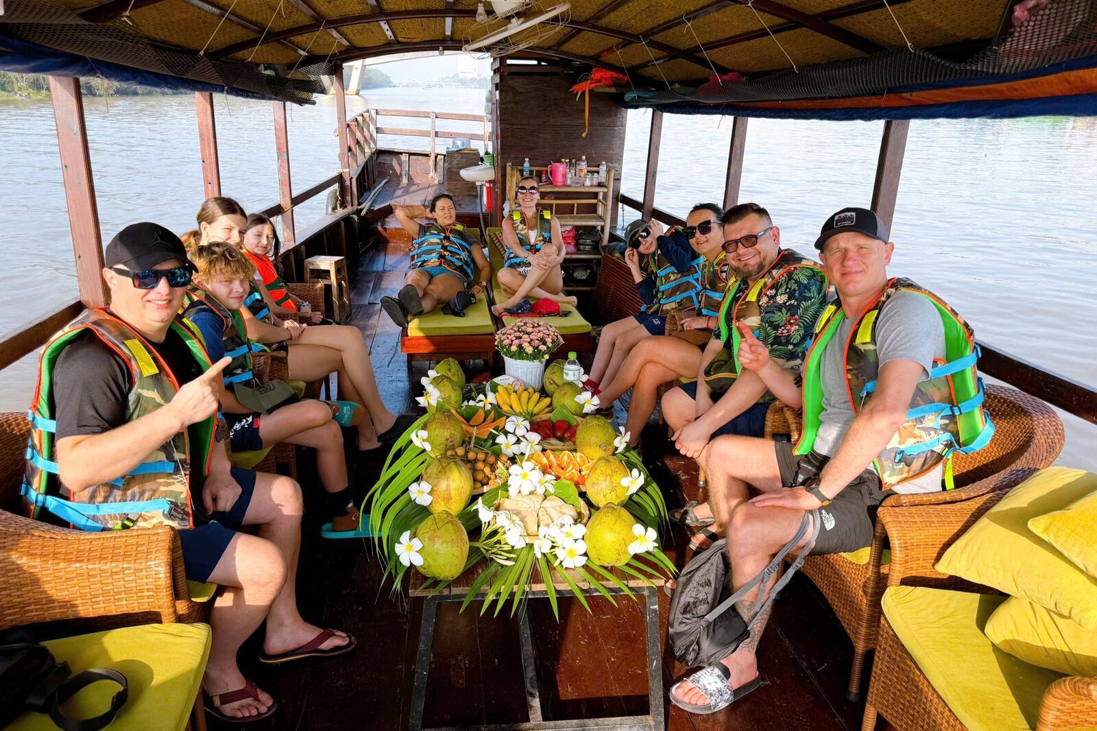 Guests in life jackets relaxing on a covered boat around a table of fresh fruit.