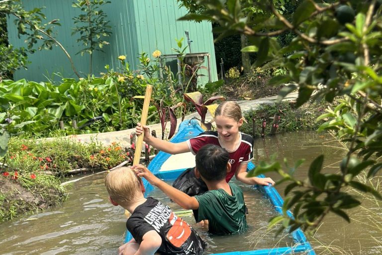Children playing with a blue boat in a shallow canal, one wading beside it.