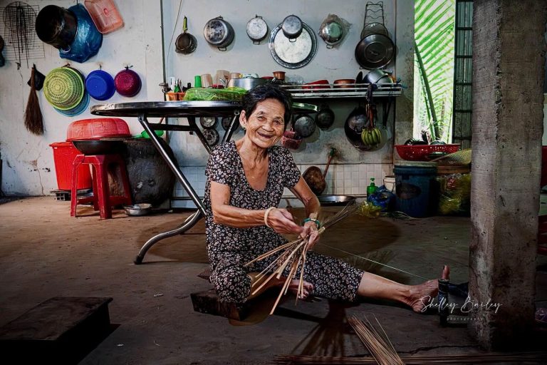 A smiling older woman sitting on a kitchen floor, binding thin stalks into a broom head.