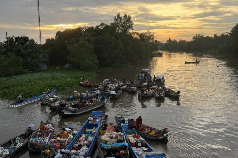 Small boats gathered on a calm river under a golden sky, trees along the banks.