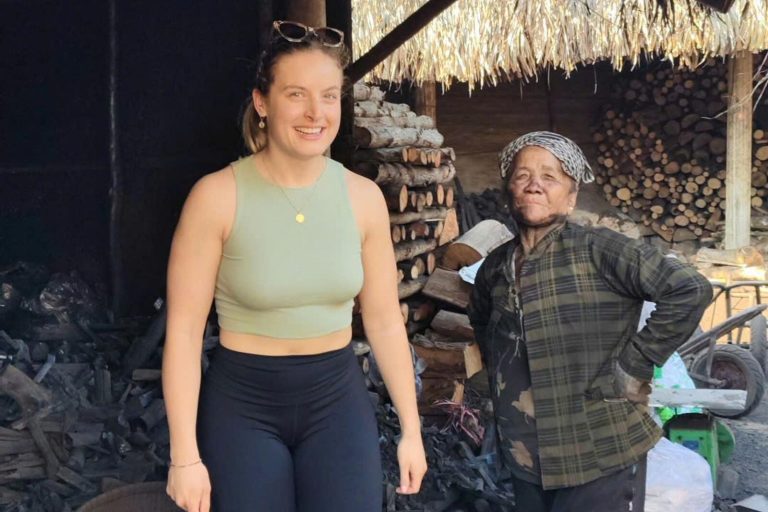 A smiling visitor beside an older local woman in front of stacked firewood.