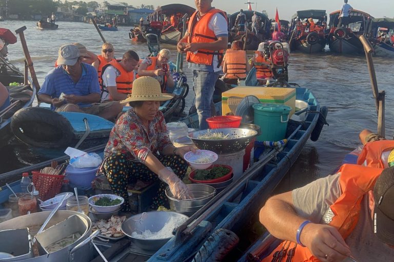 A vendor serving food from her boat to visitors in orange life jackets.