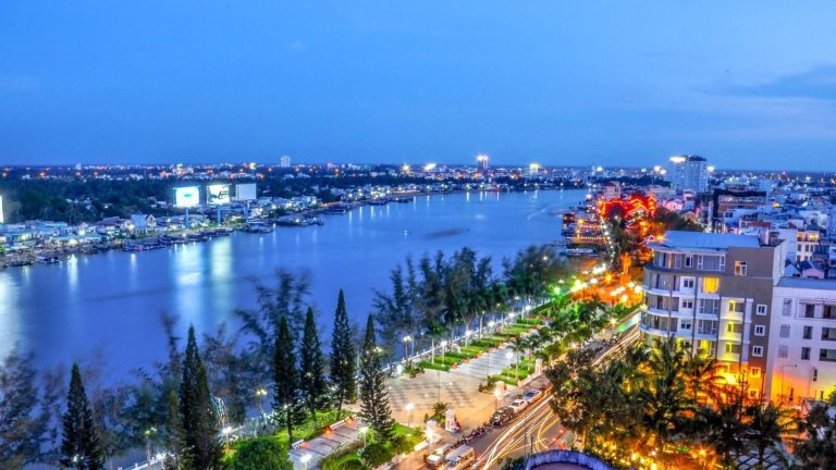 A riverside city lit up at dusk, with a park and tall buildings along the water.
