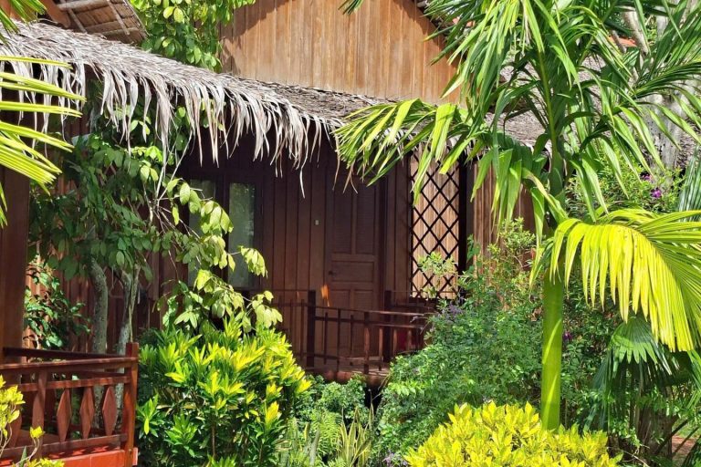 A wooden bungalow with a thatched roof among palms and leafy plants.
