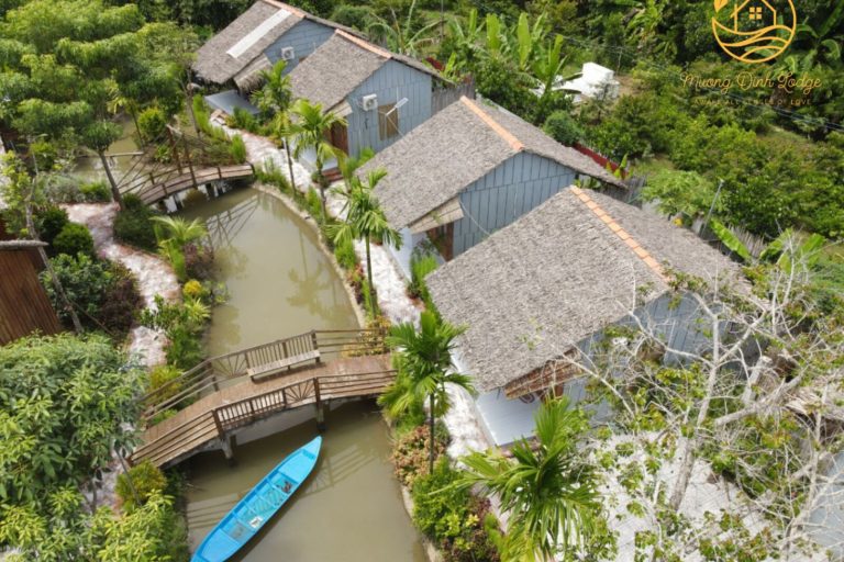 Aerial view of the lodge's thatched bungalows along a winding canal with a wooden bridge.