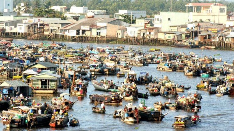 A floating market crowded with boats on a wide river below a riverside town.