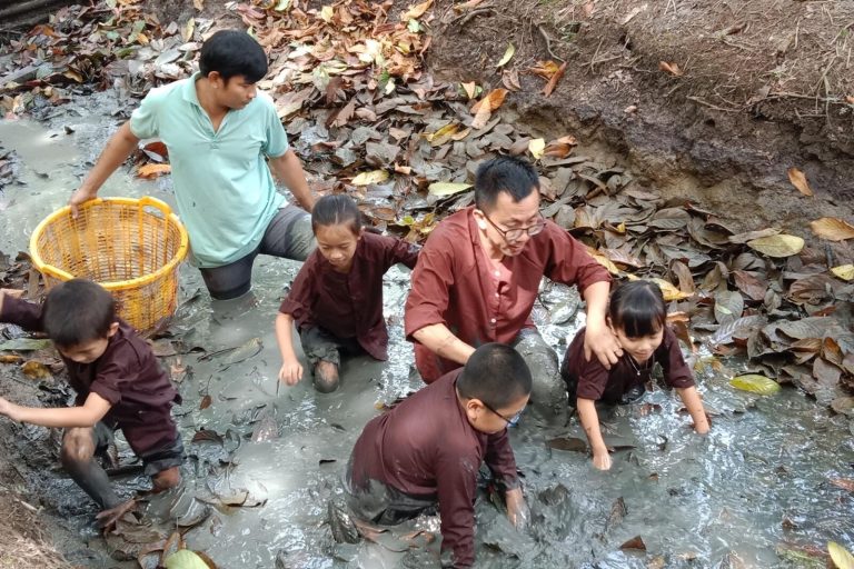 Two men and four children on their knees in a muddy ditch, one man holding a yellow plastic basket.
