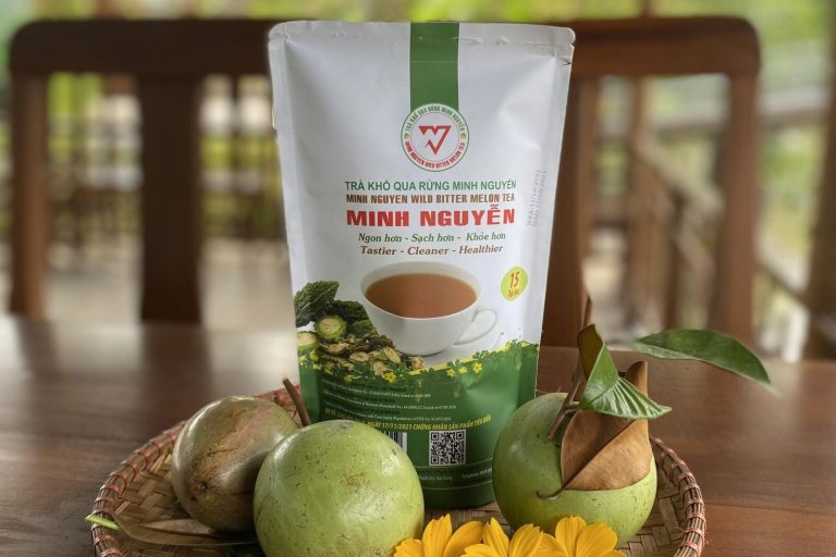 A pack of Minh Nguyen wild bitter melon tea on a table with green fruit and yellow flowers.