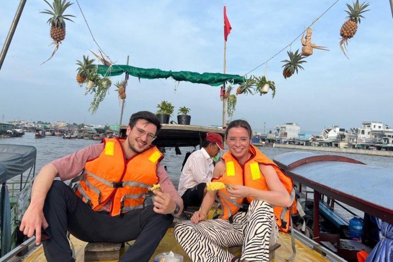 Hai du khách mặc áo phao ngồi trên ghe treo trái thơm giữa dòng sông nhộn nhịp.