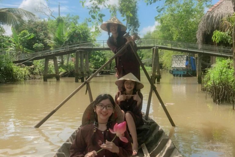 Three young women in conical hats in a wooden boat, the standing one rowing, a footbridge with metal rails behind.