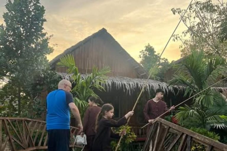 Guests holding bamboo fishing poles on a wooden bridge by a thatched cottage.