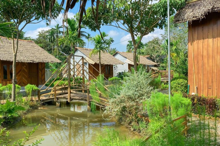 Những căn nhà mái lá và cây cầu gỗ bắc qua con mương xanh trong vườn.