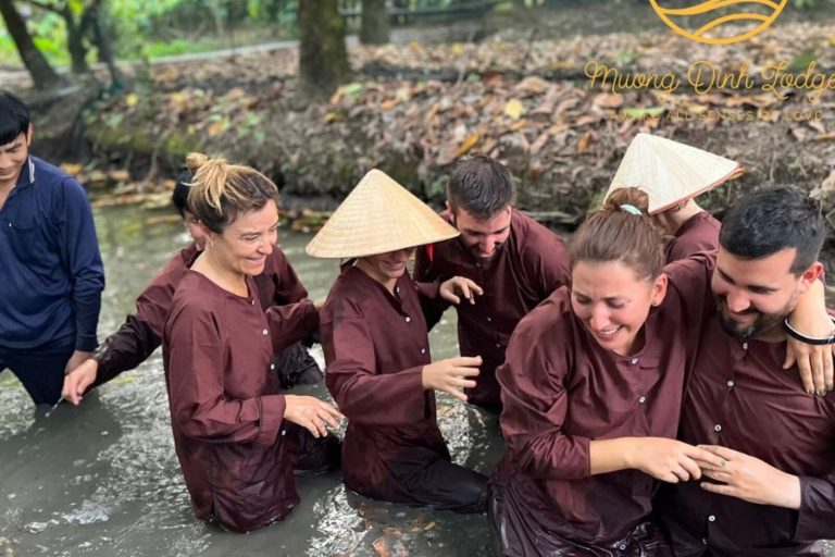 Laughing guests in brown clothes and conical hats wading in a muddy ditch, with the lodge logo.