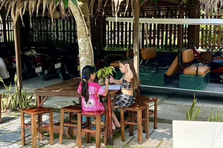 Two girls chatting at a wooden table under an open thatched shelter.
