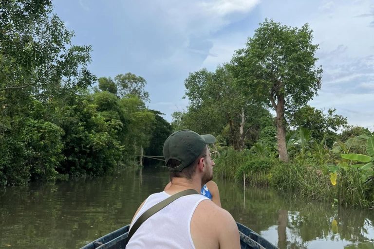 A man in a cap sitting in a boat on a quiet tree-lined canal.
