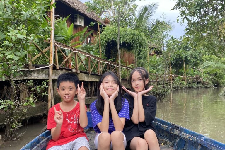 Three children making playful poses in a boat on a canal by wooden houses.